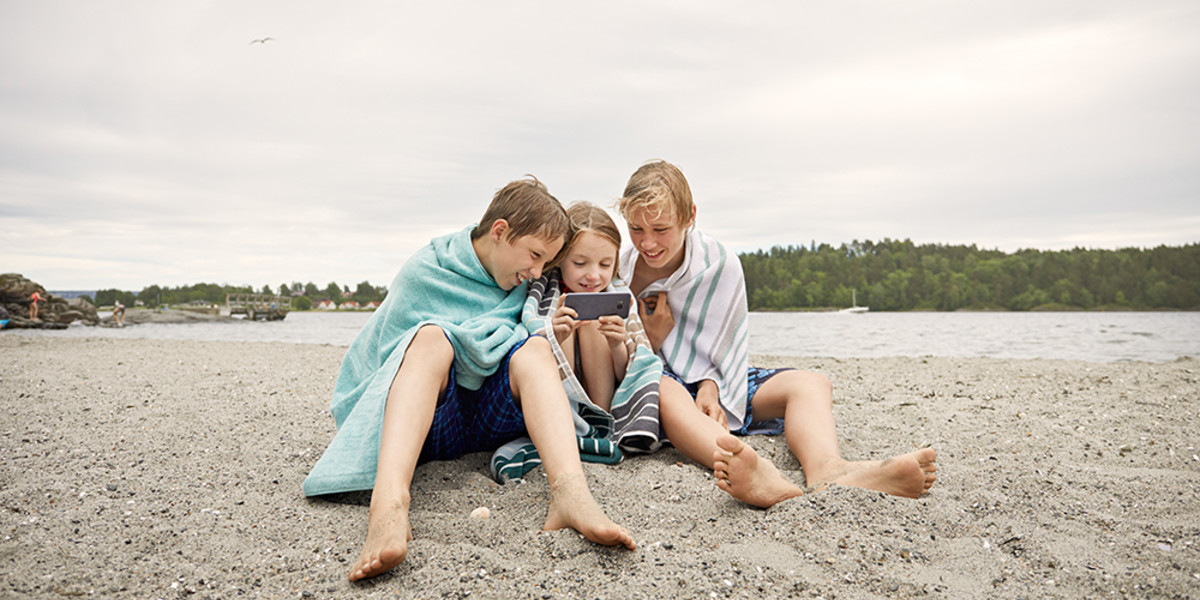 Bilde som viser tre barn på stranda hvor den midterste holder en mobil og de to andre følger med