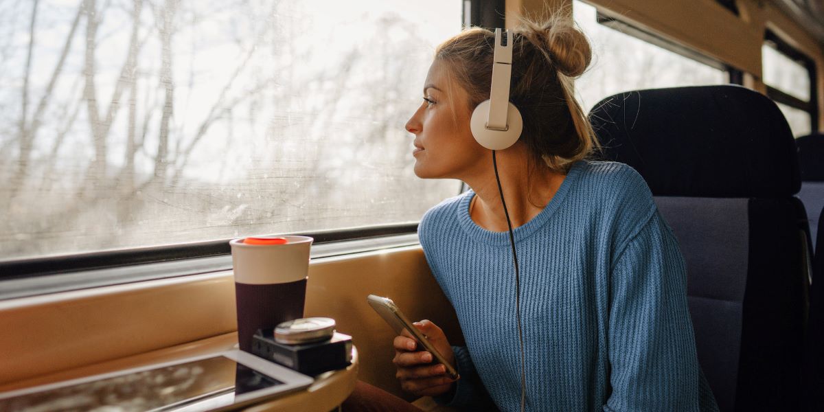 Ung kvinne sitter på toget, ser ut av vinduet og lytter til musikk på mobilen.