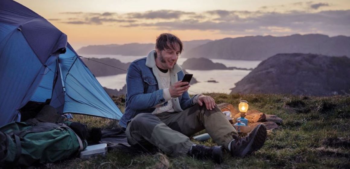 En mann sitter utenfor et telt og surfer på mobilen.