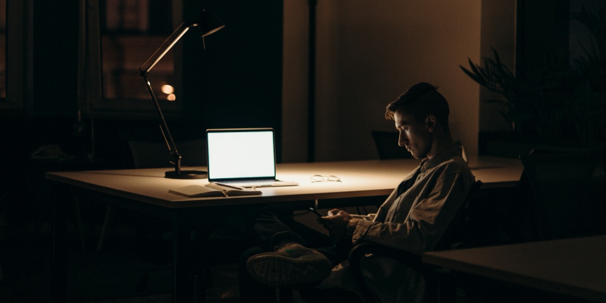 Bilde av mann som sitter ved pult i halvmørket med smarttelefon.