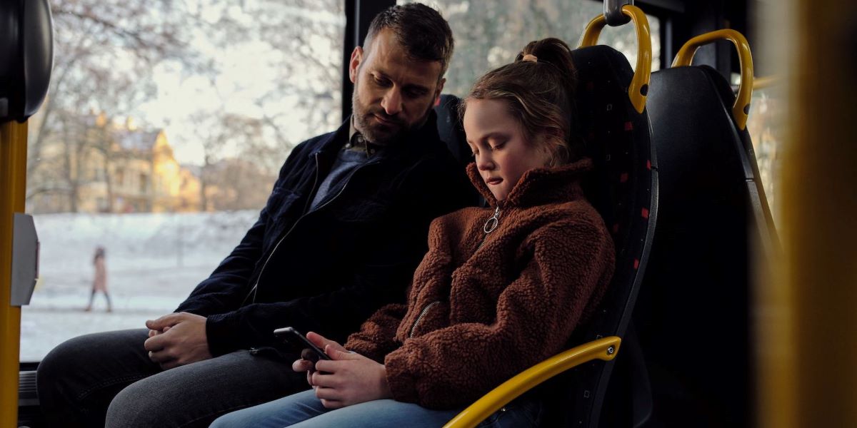 Far og datter sitter på bussen. Datteren surfer på mobilen